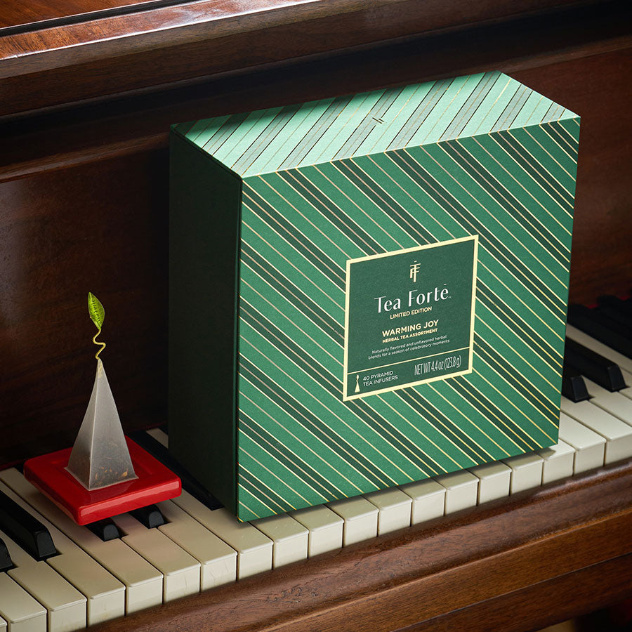 A green Tea Chest with diagonal striped pattern placed on piano keys next to a wooden piano with a tea infuser on a red tray next to it.