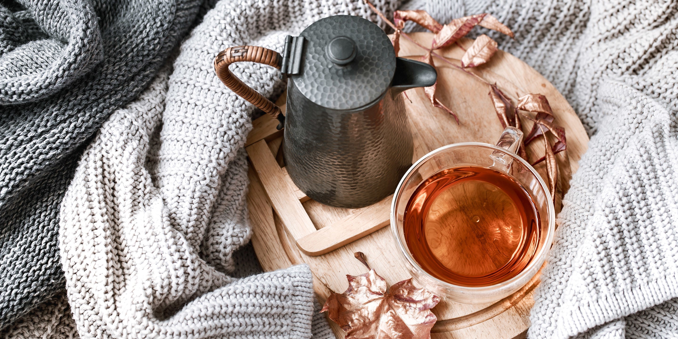 Glass cup of tea and tea kettle seen from above on a wooden tray surrounded by blankets