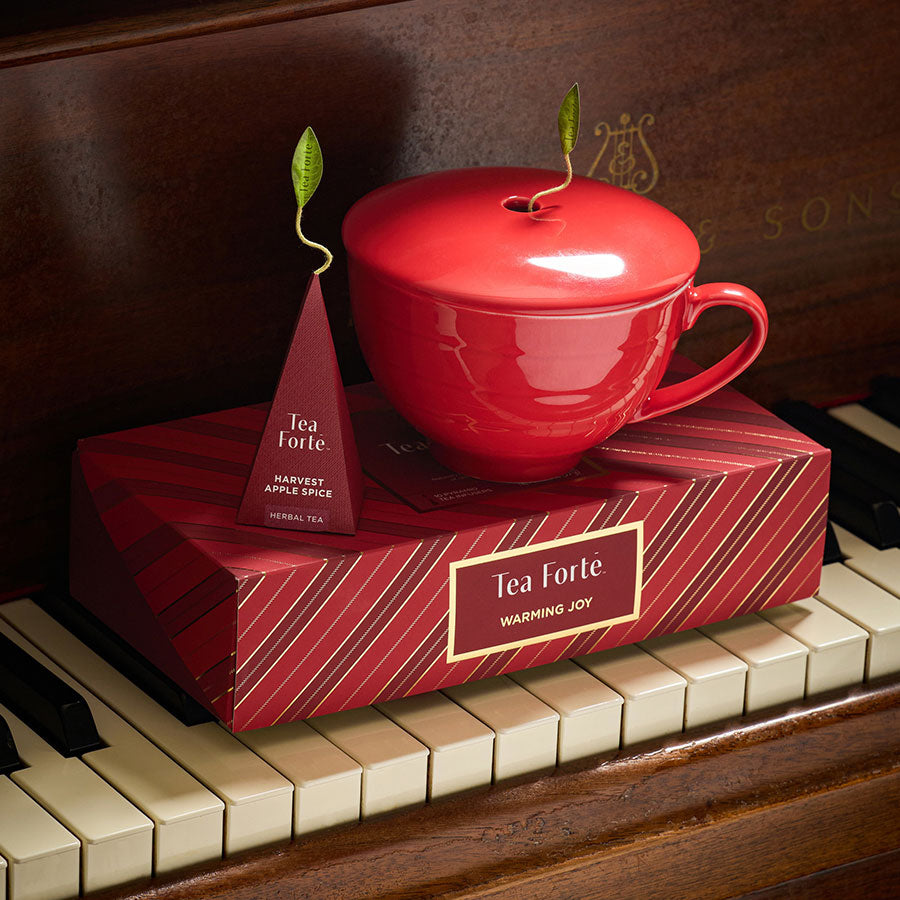 A glossy red ceramic teacup with a matching lid sits on top of a red Tea Forte Petite Presentationbox, with a tea infuser peeking out from the lid and another tea infuser standing beside it. The set is styled on a piano keyboard against a dark wood piano background.