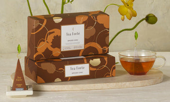 Two boxes of Tea Forte Spiced Chai tea are stacked on a marble tray next to a glass teacup filled with tea, with a single tea bag in the cup and a pyramid tea infuser on a stand in the foreground. Yellow flowers and green stems decorate the background against a light textured wall.