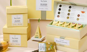 Tea tasting assortments by Tea Forte, featuring multiple boxes, including an open box showing individual pyramid tea infusers and a tea menu on the inside cover, and a small pyramid infuser on a white Tea Tray