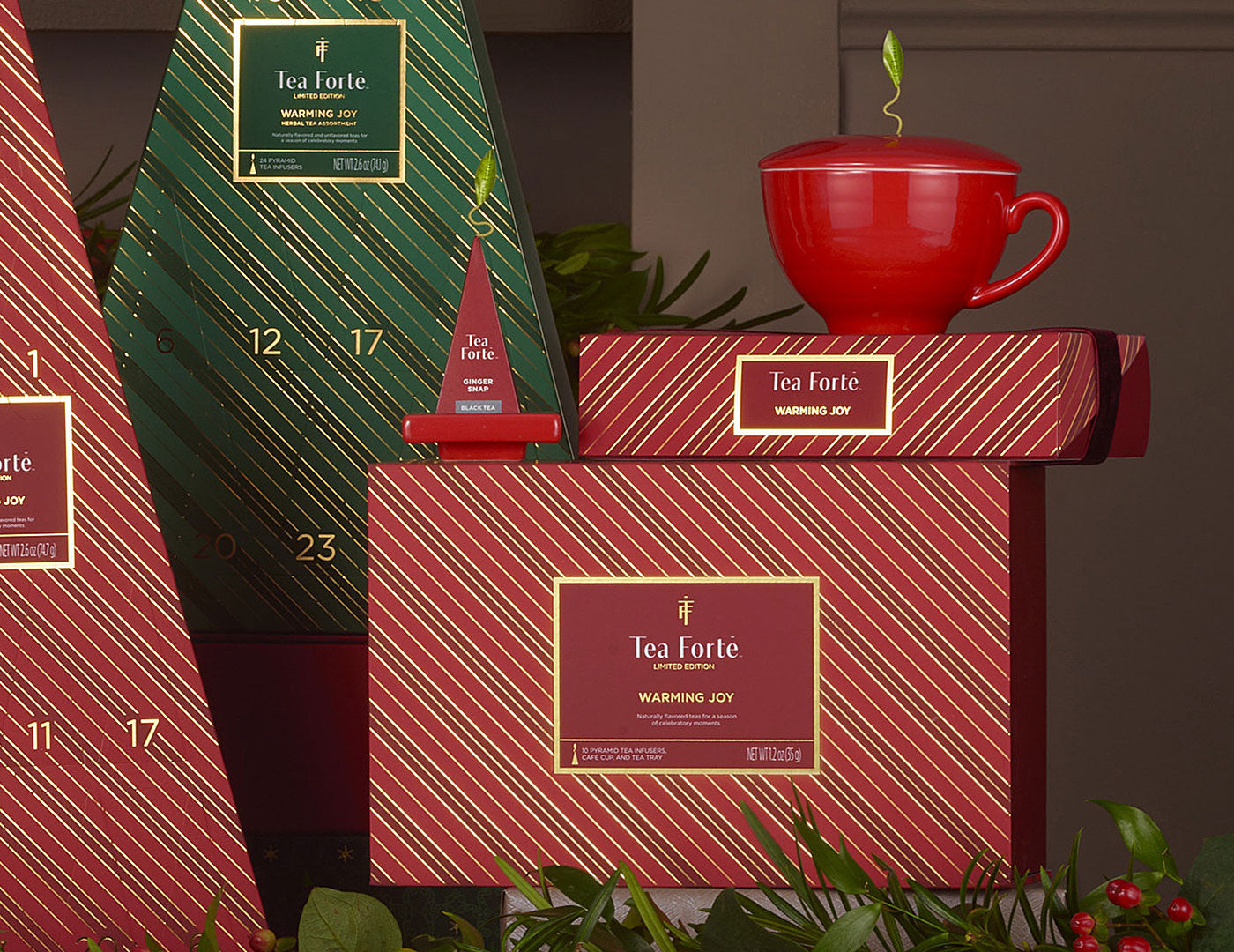 Warming Joy Gift Set styled with a red teacup and Petite BBox and Tea Tray  along with other red and green striped boxes  against a background with holiday greenery.