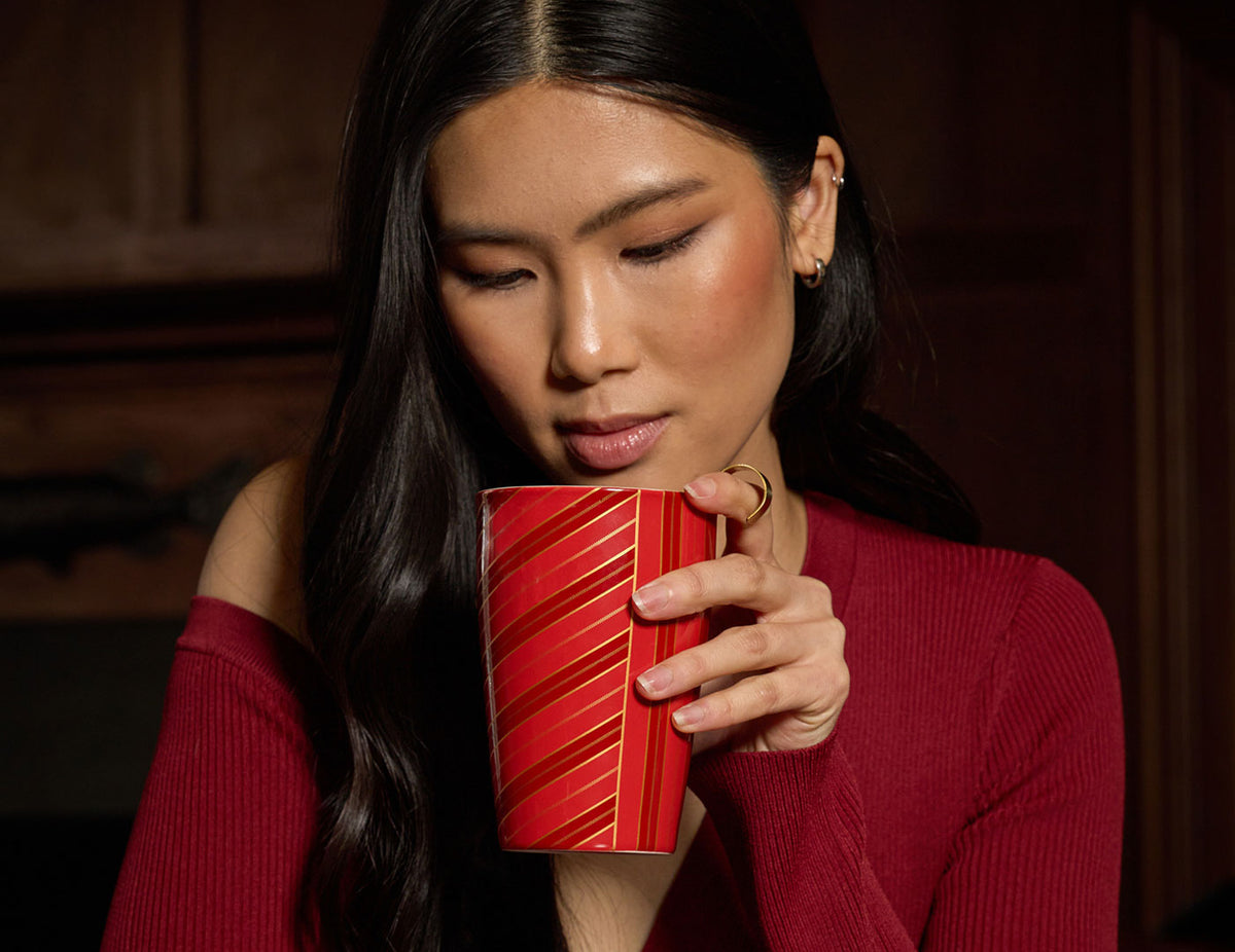 MAIN PDP | A woman with long dark hair, wearing a burgundy sweater, and a gold ring on their finger, looking at and holding a red KATI Steeping Cup Warming Joy with red and gold diagonal stripes.