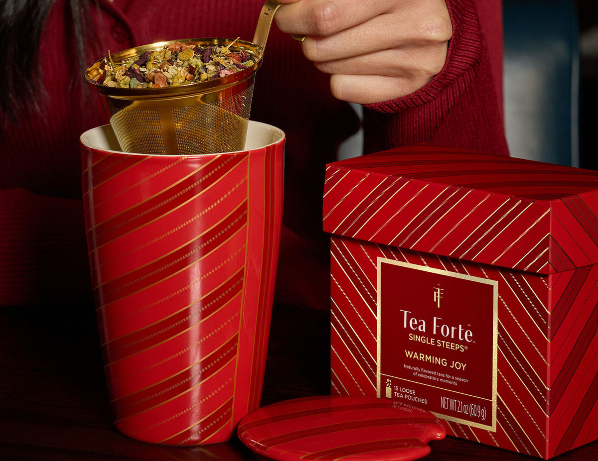 Woman holding the gold infuser basket of the Warming Joy KATI Steeping Cup with Single Steeps Sampler box to the side and lid off