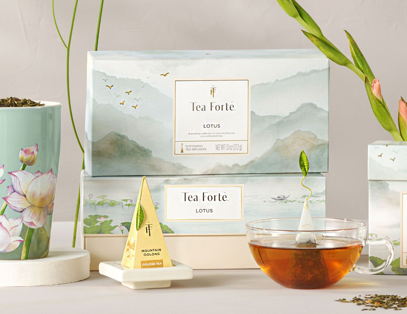 Lotus Petite Presentation Box of 10 pyramid infusers, 2 boxes stacked on top of another, with teacups ad flowers around, and infuser on white tray in front