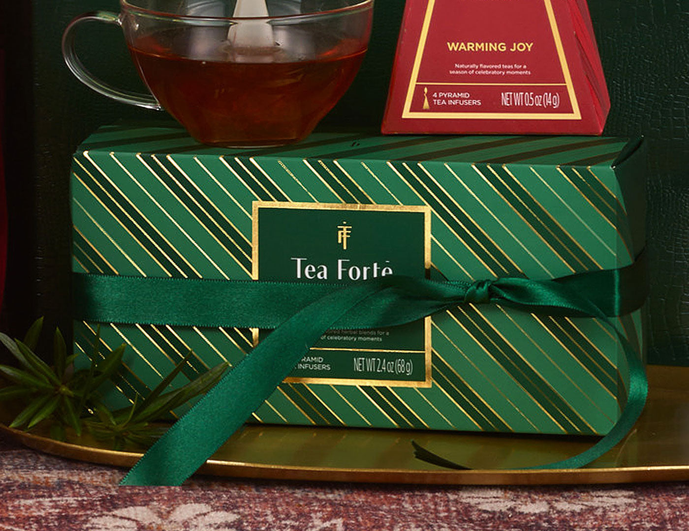 A Warming Joy Herbal Presentation Box of 20 pyrmid tea infusers, set on a gold tray, with a green ribbon tied around it, a glass cup of amber tea above, and a Warming Joy Petite Tea Tree behind it.