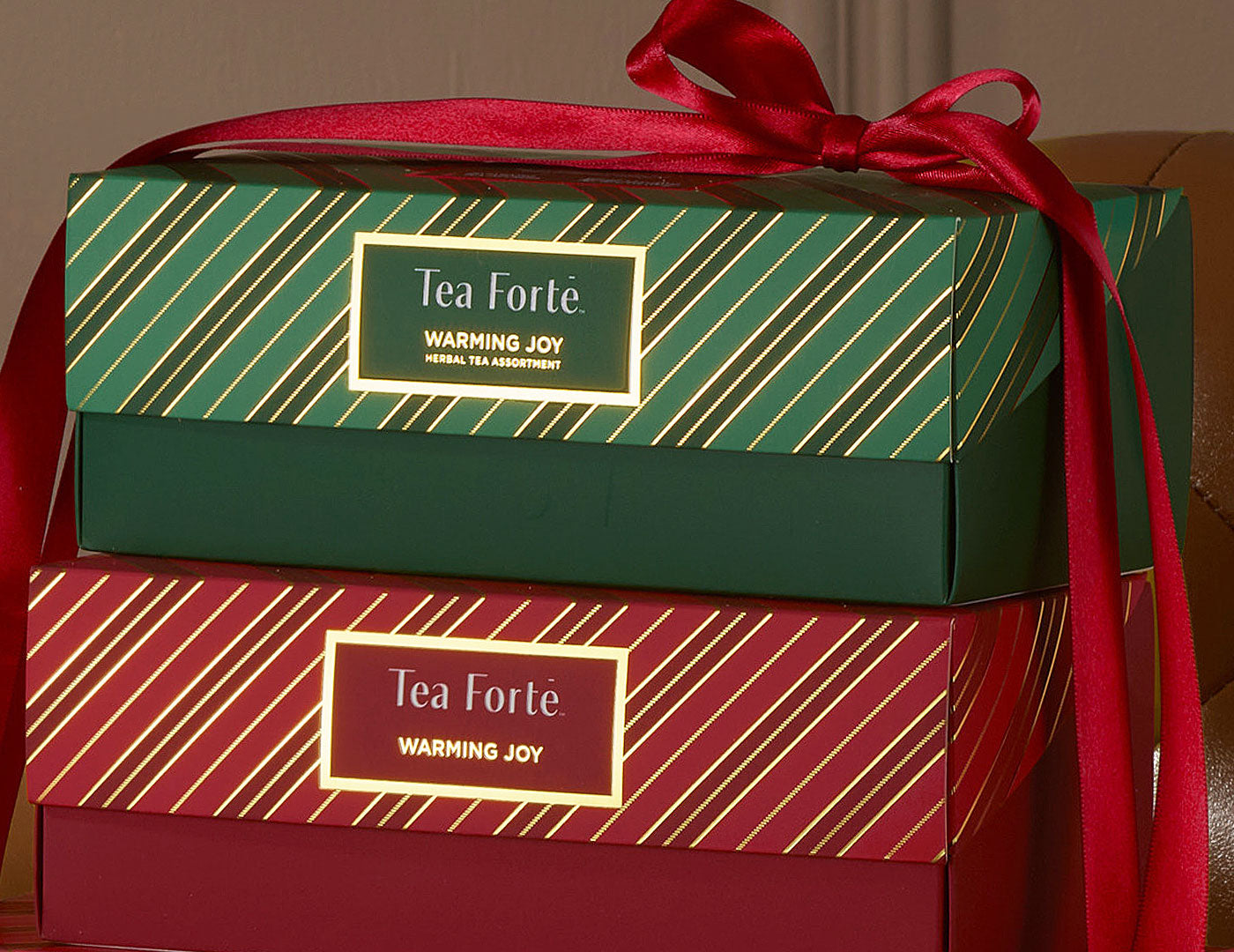 Two stacked Warming Joy Presentation Boxes with a red satin ribbon tied around both. The top box is dark green with gold diagonal stripes, and the bottom box is deep red with gold diagonal stripes.