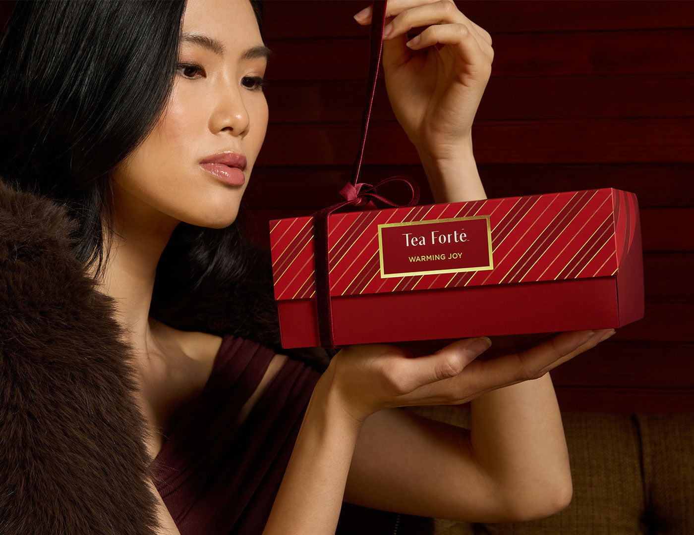 A woman with a fur shawl unties the  red ribbon around a Presentation Box Warming Joy, of 20 tea infusers, against a dark red background.