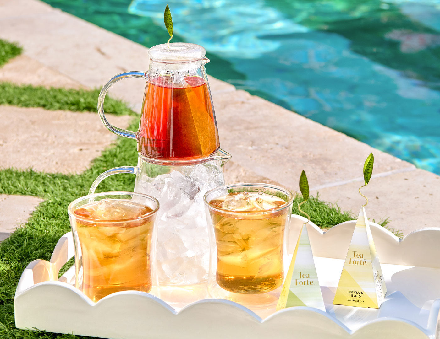 Tea Over Ice® Pitcher Set filled with iced tea and two matching POOM glasses with ice, arranged on a white tray beside a pool. Two pyramid tea infuser are visible on the tray.