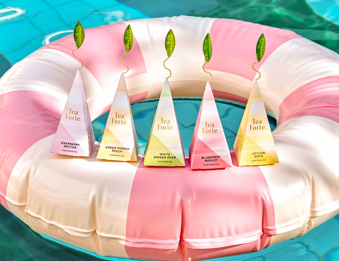 Five pyramid-shaped iced teas, each with a green leaf-shaped top, arranged on a pink and white striped inflatable pool float over turquoise water. The tea bags feature different flavor labels including Raspberry Nectar, Green Mango Peach, White Ginger Pear, Blueberry Merlot, and Ceylon Gold.