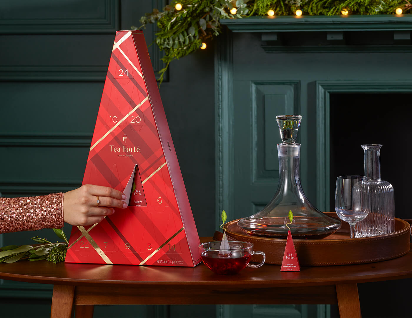 In front of a fireplace, a female hand opens a door on a Warming Joy Advent Calendar, which sits on a table with tea, a pyramid infuser a cocktail decanter and empty glasses