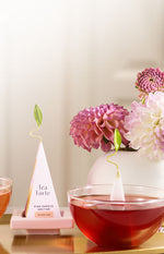 Pyramid infuser of Pink Papaya Nectar on a tea tray next to a glass cup of tea and flowers