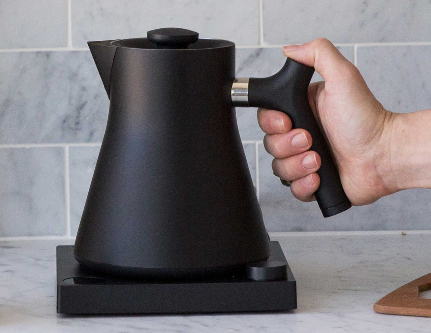 Hand on the handle of a Black Corvo Kettle EKG side view, resting on base.